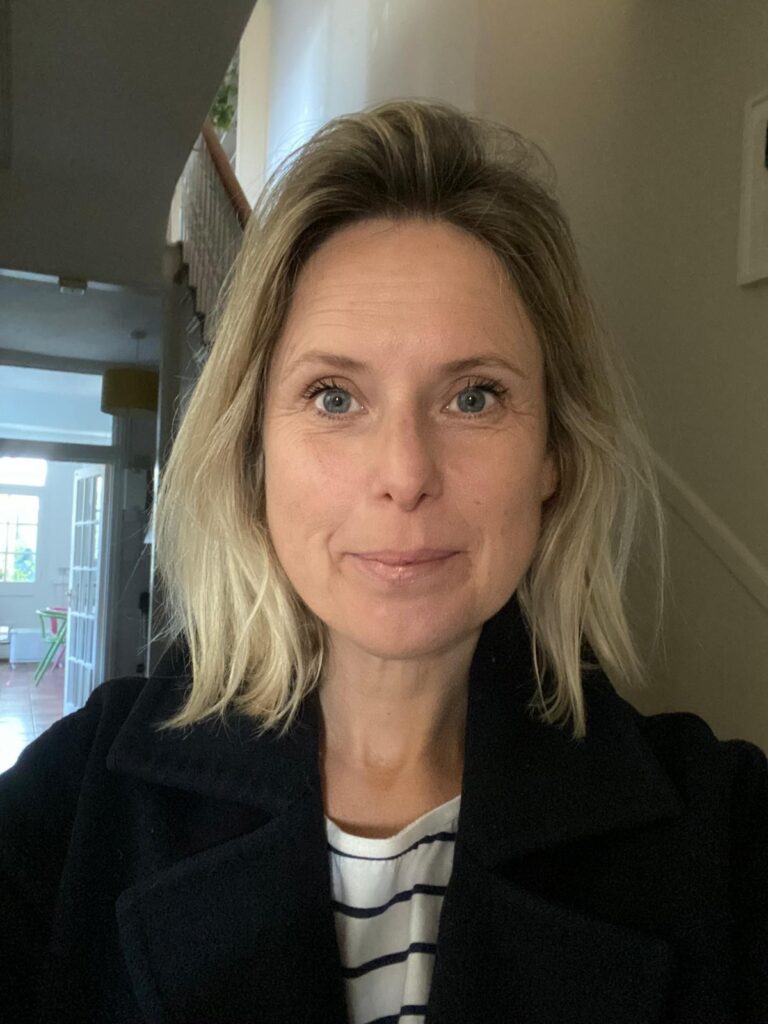 Woman with shoulder length blonde hair looking at camera, with hallway and stairs behind her